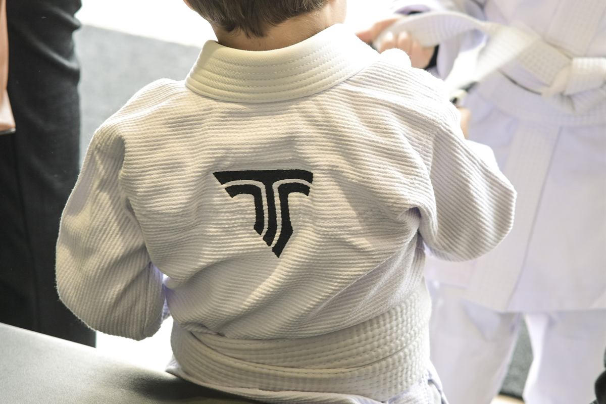 Criança pequena de costas, com o kimono da Trivium, tendo a faixa amarrada por um adulto